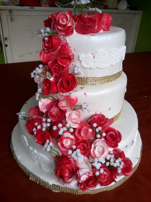 A white wedding cake decorated with red flowers and golden ribbon balls, baked by NHK Home Bakery